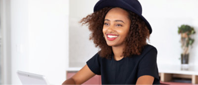 En kvinna med lockigt hår i en mörkblå mössa och svart skjorta ler när hon sitter vid ett bord med en bärbar dator.