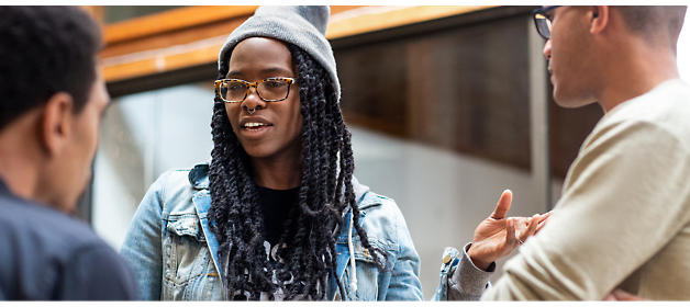Person with long braided hair and glasses wearing a gray beanie and denim jacket is speaking to two others indoors.