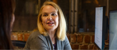 Kvinna med hår som sitter vid ett skrivbord och pratar med en annan person. Två datorskärmar visas bredvid hennes