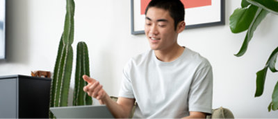 En person i en ljusgrå tröja sitter inomhus, ler och gestikulerar när han använder en bärbar dator