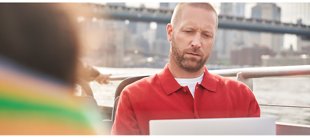A man with a beard and wearing a red shirt is focused on a laptop while sitting on a boat with a bridge 