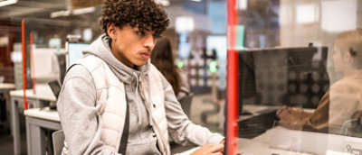 A person with curly hair, wearing a gray hoodie and white vest, works on a computer in a library or workspace