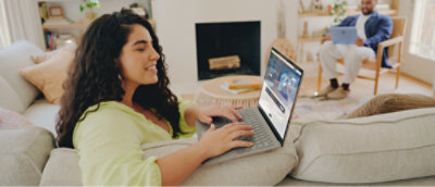 Una mujer con el pelo largo y rizado utiliza un portátil sentada en un sofá. Un hombre sentado en el fondo usando un portátil