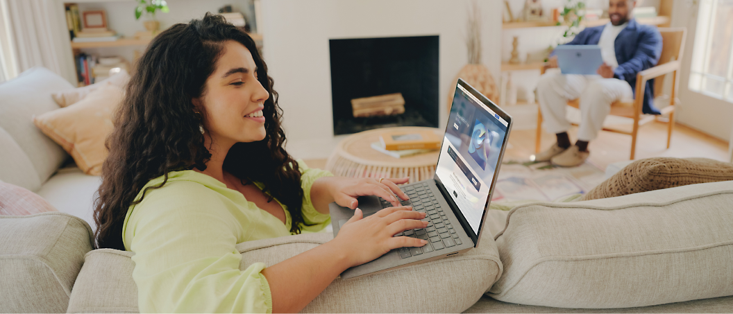 A woman with long curly hair uses a laptop while sitting on a couch. A man sits in the background using a laptop