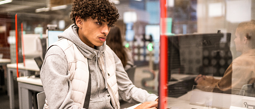 A person with curly hair, wearing a gray hoodie and white vest, works on a computer in a library or workspace