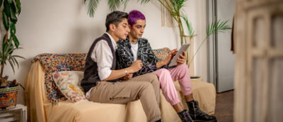 Two people sitting together on a couch, looking at a tablet. One has short gray hair and wears beige pants