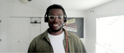 白い壁と窓のある明るい部屋で、眼鏡とヘッドホンを身に着けた人物が笑っている 