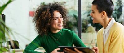 Two women, one with curly hair, smiling and looking at a tablet together in a brightly lit office space.