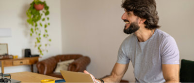 A man with a beard and striped shirt smiles while holding a tablet in a cozy room with plants and a couch in the background.