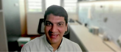 短い黒髪の人物がカメラに向かって笑っている。背景がぼやけた明るい部屋に座っている