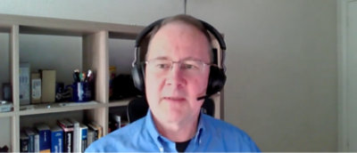 ヘッドホンとマイクを装着している人物が、本やさまざまなアイテムでいっぱいの本棚の前に座っている。