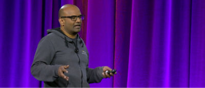 暗い色のパーカーを着て眼鏡をかけている人物が、背景に紫色のカーテンがかかっているステージでヘッドセット マイクに向かって話している