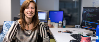 オフィスのデスクに座っている女性。周囲にはコードが表示されているコンピューター画面、コーヒー カップ、オフィス用品がある