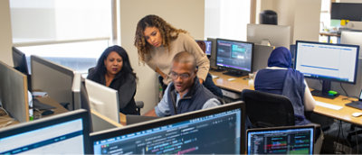 4 人の人物 (1 人はヒジャーブを着用) がオフィス環境でコンピューターに向かって仕事をしている。1 人の人物は立っている 