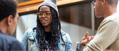 Three young adults engaged in conversation outdoors, with a woman in a denim jacket and glasses 