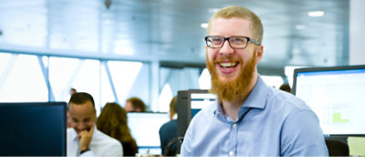 同僚と慌ただしいオフィス環境でデスクに座りながら、赤いひげを生やして眼鏡をかけている男性が笑顔を見せている 