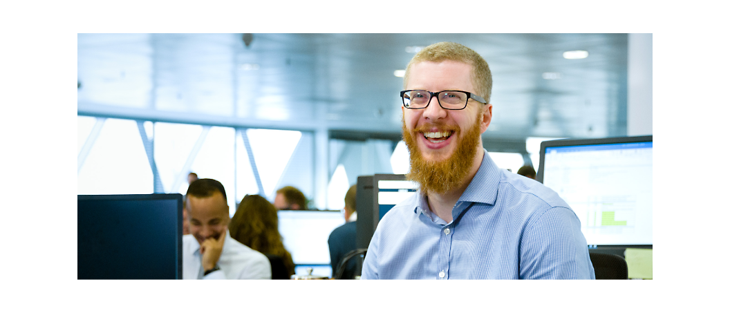A man with a red beard and glasses smiles while sitting at his desk in a busy office environment with colleagues 