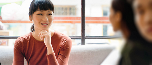 一名亞洲女性身穿紅色上衣、手托下巴微笑，坐在明亮的咖啡廳中