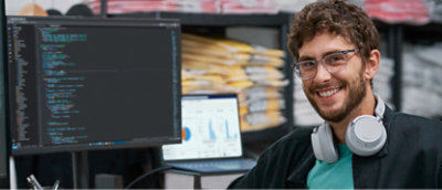 眼鏡をかけてヘッドホンを首回りにかけている男性が、コードが表示されているコンピューター画面とグラフが表示されてるノート PC の前で笑っている。