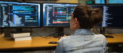 明るいオフィス環境でコードが表示されている複数のコンピューター画面に向かって仕事をしている、デニム ジャケットを着た人物。