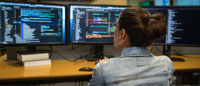 Person in a denim jacket working on multiple computer screens displaying code in a well-lit office environment.