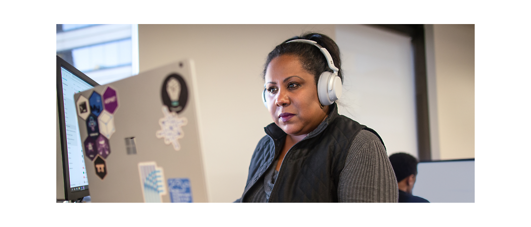 A woman wearing headphones works on a computer in an office. The laptop has several stickers on the back.