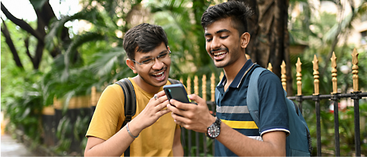 Two young men laughing and looking at a smartphone together, standing outdoors with greenery in the background.