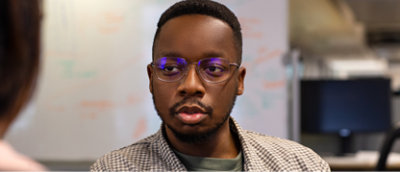 眼鏡をかけてチェック柄のシャツを着ている人物が、背面がカメラに向いている別の人物の方を見ている。