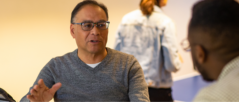 Man in glasses and grey sweater having a conversation with another person. A third person is seen in the background,
