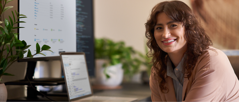 A person with curly hair smiles at the camera while working on a laptop and a large monitor displaying code