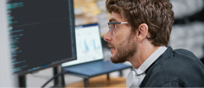 ひげを生やして眼鏡をかけ、ヘッドホンを首にかけている人物が、表示されているコンピューターの画面を見ている 