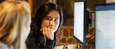 Two women in an office setting, one asian and one caucasian, focusing on a computer screen visible in the background.