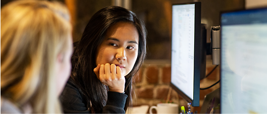 Dos mujeres en una oficina, una asiática y otra caucásica, se concentran en la pantalla de un ordenador que se ve al fondo.