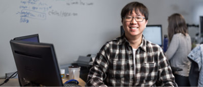 A smiling person with glasses wearing a plaid shirt, sitting at a desk with a computer monitor, in an office with a whiteboard