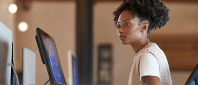 A young woman working attentively at a standing desk with computer monitors in a modern office setting.