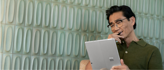 一名戴眼​​鏡身穿綠色襯衫的中年亞洲男子，沉思地看著靠在帶有紋理的綠牆上的平板電腦 