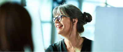 Smiling middle-aged woman with glasses and a bun discussing with a colleague in a bright office setting.