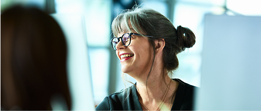 Mulher de meia-idade sorrindo, com óculos e um coque, discutindo com um colega em um ambiente de escritório iluminado.