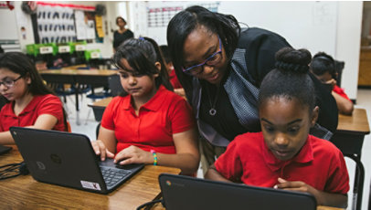 A teacher watches young girls work on laptops.
