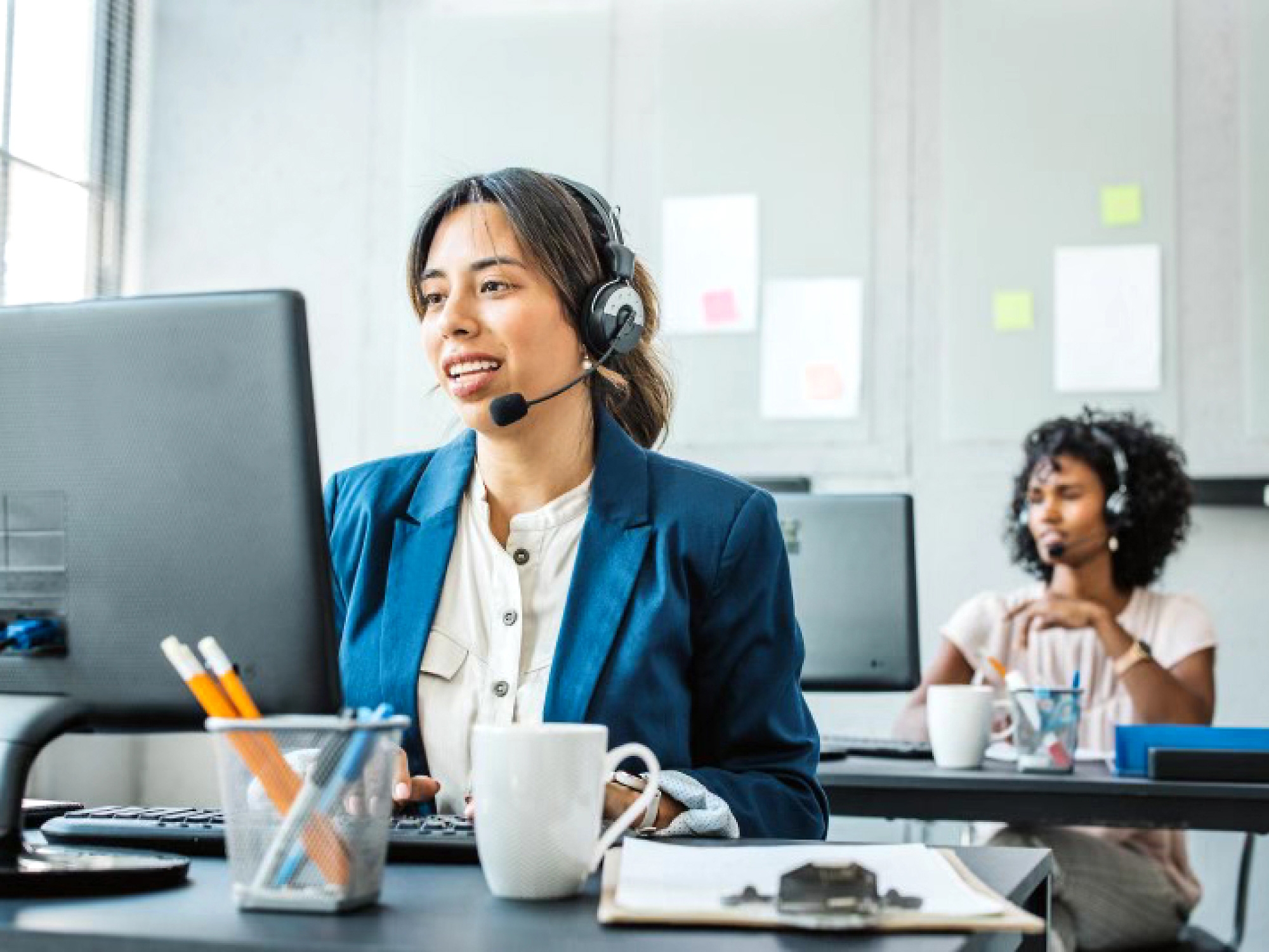Een medewerker van de klantenservice die een headset draagt en glimlacht terwijl ze op haar computer in een kantooromgeving werkt.