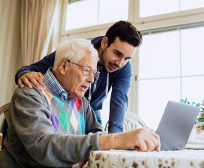 Young man assists elderly man using a laptop at a table near a window.