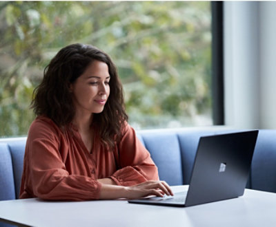 A woman in a rust-colored blouse working on a laptop at a white table near a window with a leafy view.