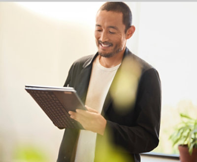 Man in a black jacket smiling and reading a document in a sunlit room.