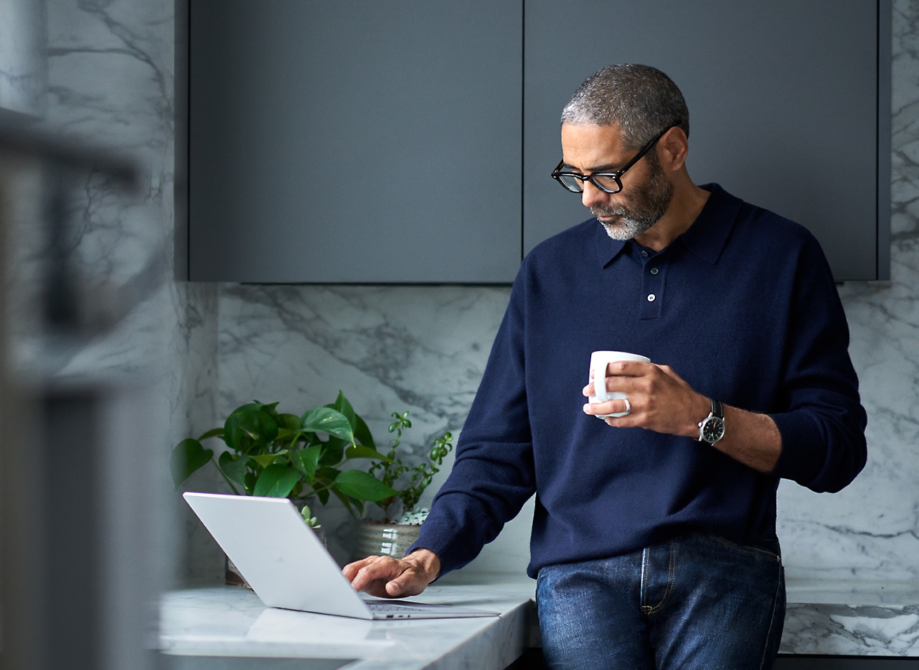 Middle-aged man with glasses, holding a cup and using a laptop on a marble counter in a modern kitchen.