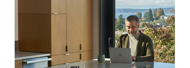 A man working on a laptop at a sleek kitchen counter with a scenic lake view in the background.