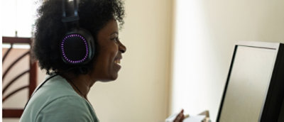 A middle-aged black woman wearing illuminated headphones works intently at a computer in a home office setting.