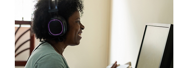 A middle-aged black woman wearing illuminated headphones works intently at a computer in a home office setting.