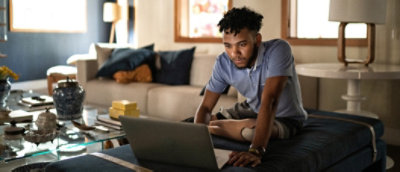 A man working on a laptop on the couch in a well-lit living room, looking intently at the screen.