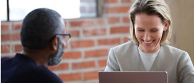 Two professionals, a woman and a man, are smiling and discussing over a laptop in a modern office setting.
