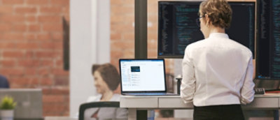 A professional woman standing at a standing desk, reviewing code on multiple computer screens